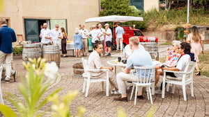 Gemeinsamer Moment im Innenhof des Weingut Robert König: Unter einem großen Sonnensegel versammeln sich die Gäste, während an den weißen Tischen Wein, Gläser und sommerliche Leckereien bereitstehen.