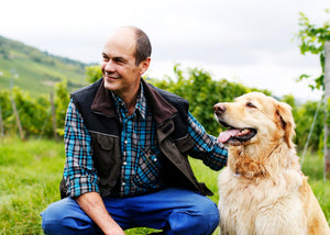 Winzer Robert König der 2 kniet im grünen Weinberg in Rüdesheim und blickt über die Rebstöcke seines Weinguts, während er sanft seinen Hund streichelt. Im Hintergrund erstrecken sich gepflegte Reben und die hügeligen Rüdesheimer Weinberge, die eine eindrucksvolle Weinlandschaft bilden.